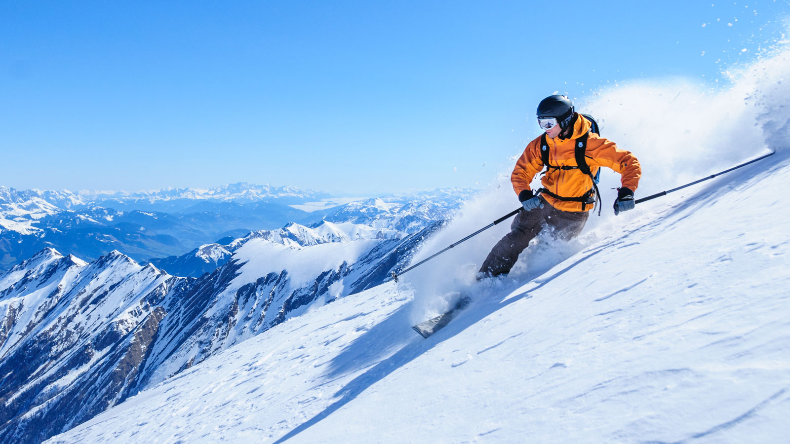 Skier in bright orange jacket making a sharp turn on a steep snow-covered slope with a spectacular panoramic view of alpine peaks in the background, highlighting an action-packed skiing adventure.